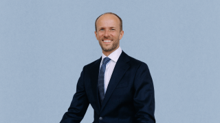 Bogumil Baranowski, dressed in a dark suit, white shirt, and patterned tie, stands and smiles in front of a plain light blue background.
