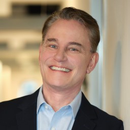 Smiling man with short brown hair wearing a dark blazer and light blue shirt, standing indoors against a light-colored wall.