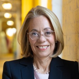 A woman with blonde hair and glasses, wearing a black blazer and a pearl necklace, stands indoors in front of a yellow wall, smiling at the camera.