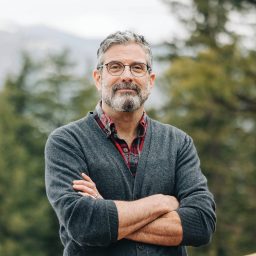 Man with gray hair and beard wearing glasses and a cardigan stands outdoors with arms crossed, trees and mountains in the background.