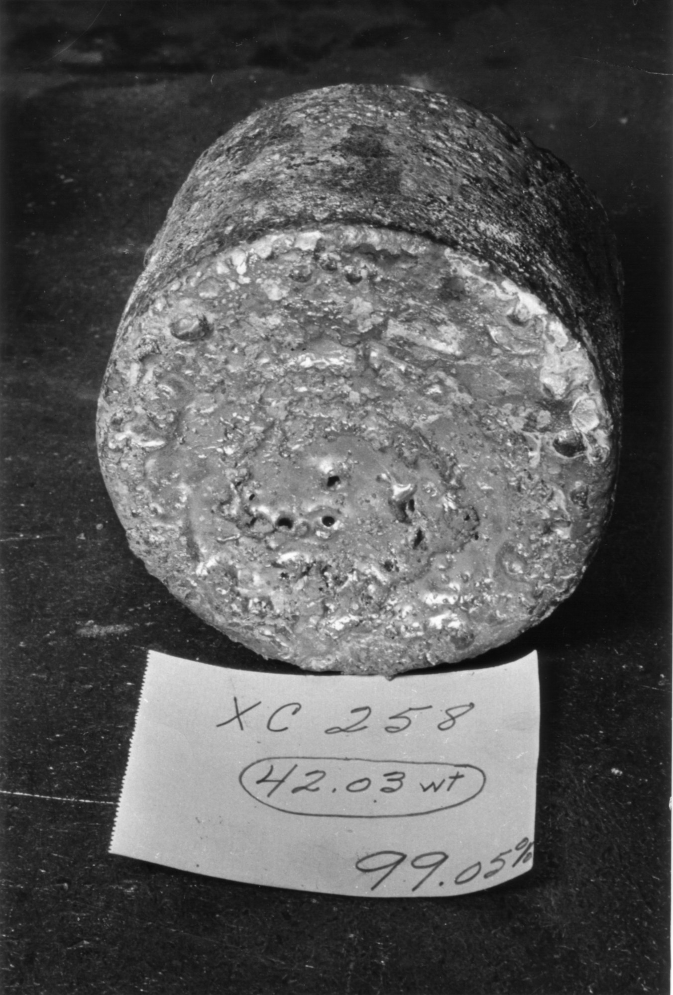 A cylindrical metal ingot sits on a dark surface next to a handwritten note reading “XC 258, 42.03 wt, 99.05%.”.