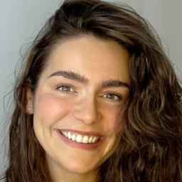 A woman with long wavy brown hair and thick eyebrows is smiling at the camera against a plain light gray background.