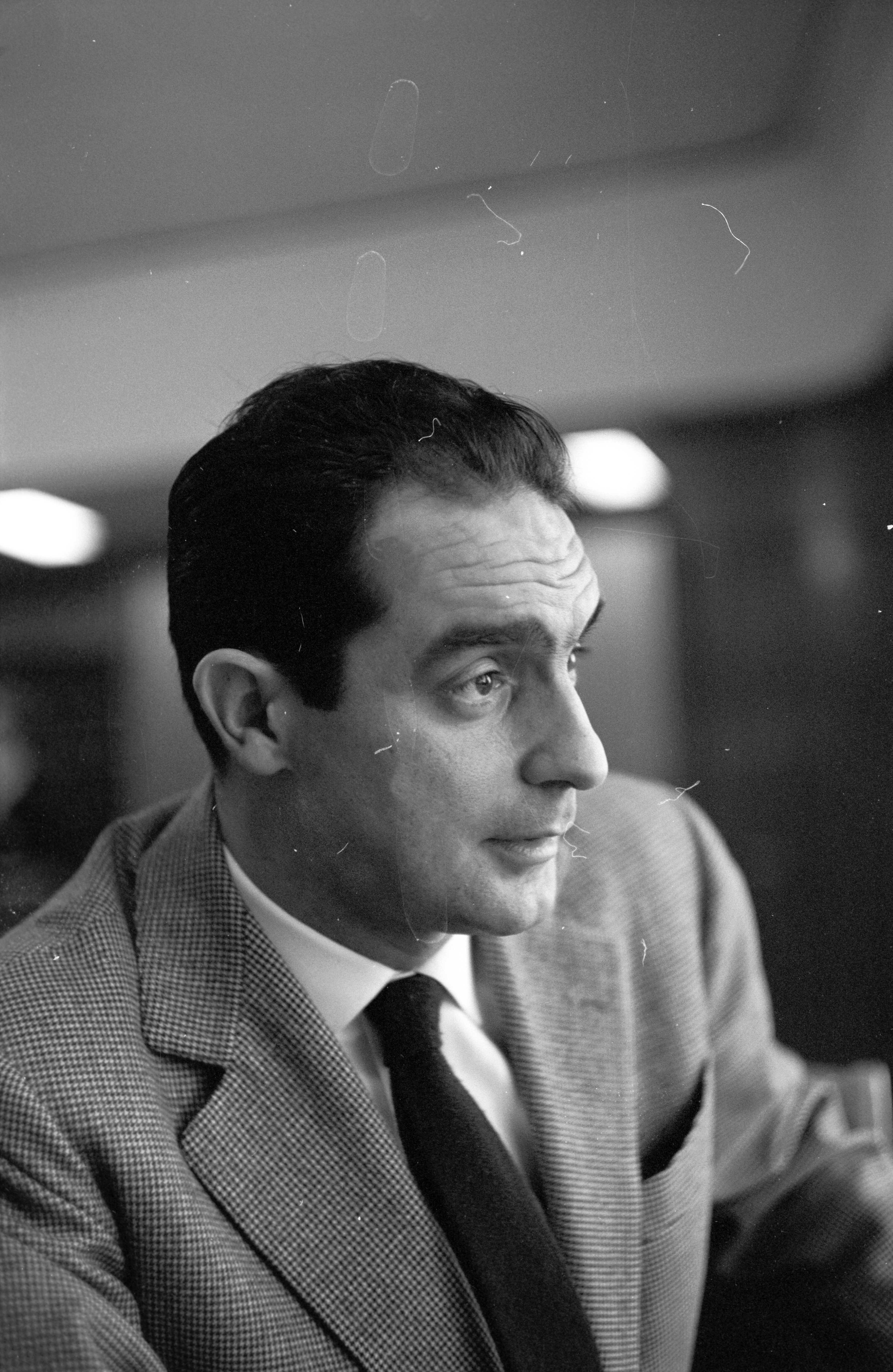 A man in a suit and tie is looking to the side in a black and white photo, with a blurred indoor background.