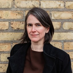 A woman with short brown and gray hair wearing a black coat stands in front of a yellow brick wall, looking at the camera.