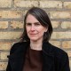 A woman with short brown and gray hair wearing a black coat stands in front of a yellow brick wall, looking at the camera.