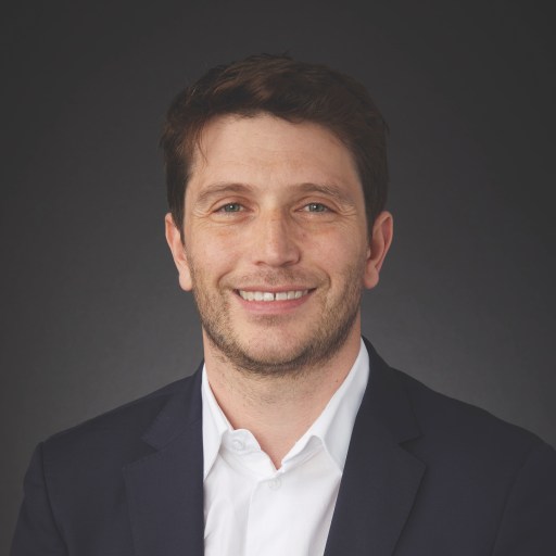 A man with short brown hair and a trimmed beard, wearing a white shirt and dark blazer, smiles at the camera against a plain dark background.