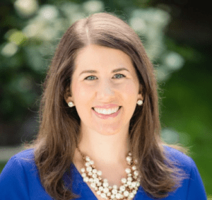 Woman with long brown hair, wearing a blue top, pearl necklace, and earrings, smiling outdoors with greenery in the background.