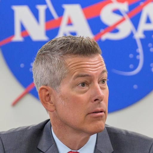 A man in a suit sits in front of a large NASA logo sign, symbolizing the determination to defy charter limits and push the boundaries of space exploration.