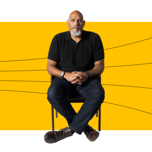 A man with a bald head and beard sits on a chair with arms resting on his lap, in front of a yellow rectangle with black lines and dots extending outward.