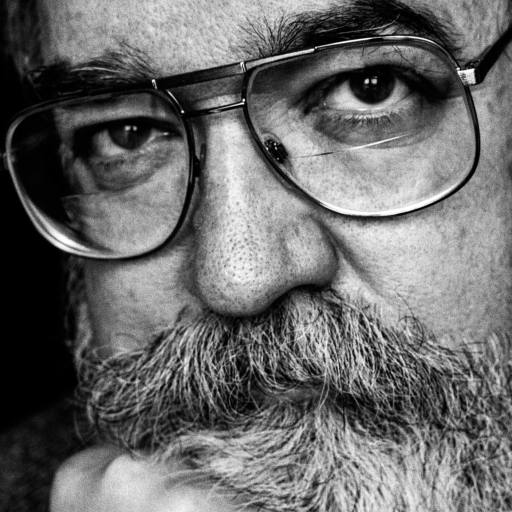 Black and white close-up of an older man with glasses, a bushy mustache, and beard, resting his chin on his hand and looking directly at the camera.