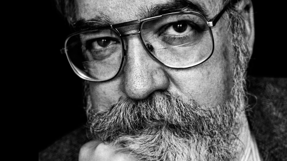 Black and white close-up of an older man with glasses, a bushy mustache, and beard, resting his chin on his hand and looking directly at the camera.