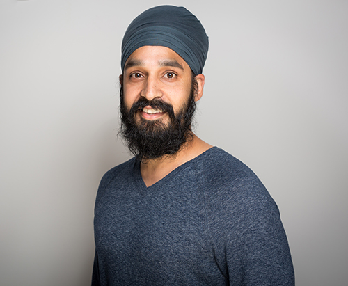 Man with a dark beard and mustache wearing a blue turban and blue sweater, standing against a plain light gray background, looking at the camera and smiling.