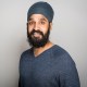 Man with a dark beard and mustache wearing a blue turban and blue sweater, standing against a plain light gray background, looking at the camera and smiling.