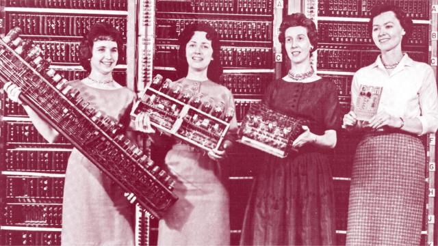 Four women stand in front of a vintage computer, each holding different components of its hardware, with the computer's panel visible in the background—an image reminiscent of historical innovation studied by Angus Fletcher.