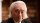 An older man with long white hair and a suit looks at the camera, standing in front of a blurred background with bookshelves.