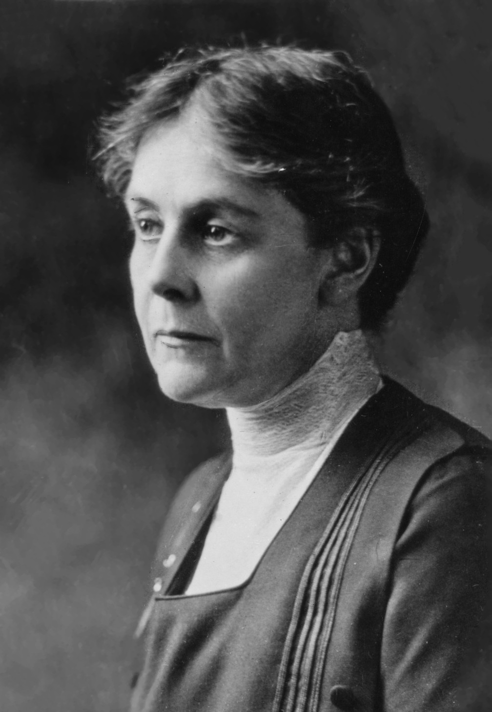 Black and white portrait of a woman with short wavy hair, wearing a high-collared blouse and dark dress, facing left with a neutral expression.