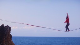 A person walks barefoot on a slackline stretched high above the ocean, with one arm raised for balance and cliffs visible at each end.
