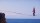 A person walks barefoot on a slackline stretched high above the ocean, with one arm raised for balance and cliffs visible at each end.