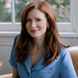 A woman with long red hair wearing a blue button-up shirt sits on a tan chair in front of a window with greenery outside.