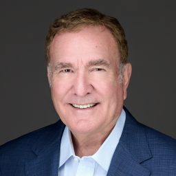 Smiling older man with short light brown hair wearing a blue blazer and light blue collared shirt against a plain dark background.