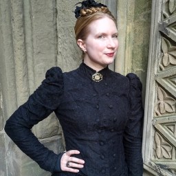 A woman wearing a high-necked, black Victorian-style dress poses near a stone wall with architectural details.