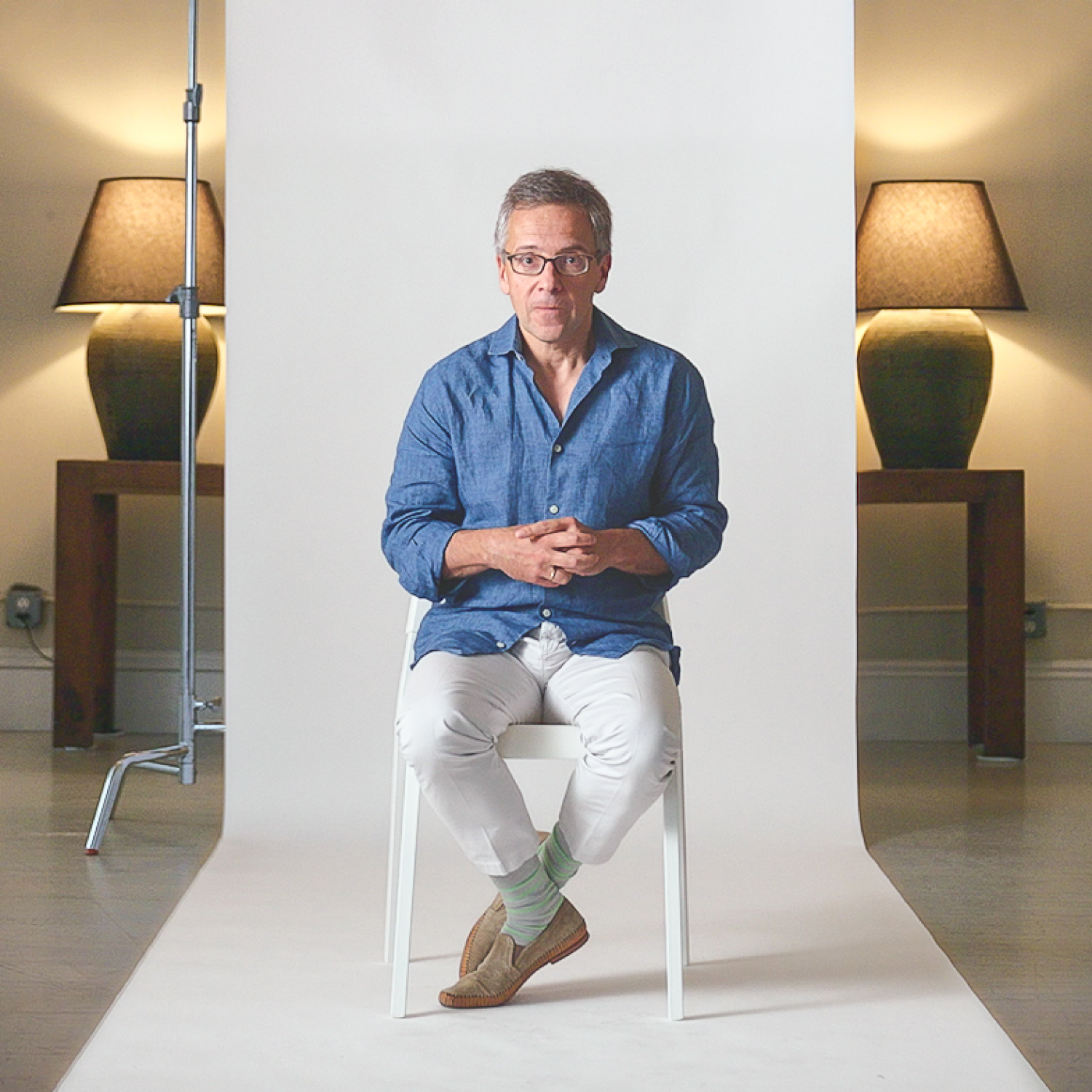 A man in a blue shirt and white pants sits on a white chair in a studio, with lamps and tables in the background and a white backdrop behind him.