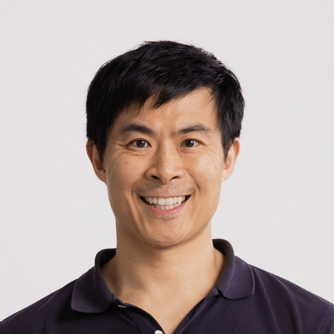 A man with short black hair, wearing a dark collared shirt, smiling at the camera against a plain light background.