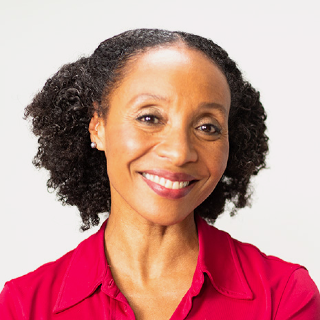 A woman with curly black hair, wearing a red collared shirt, smiles at the camera against a plain white background.
