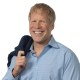 Smiling man with blond hair in a light blue button-down shirt holds a dark jacket over his shoulder against a white background.