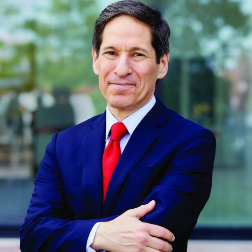 A man in a navy suit and red tie stands outdoors with arms crossed, smiling slightly at the camera.
