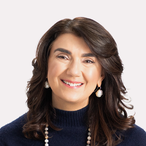 A woman with medium-length brown hair, wearing a navy sweater, pearl earrings, and a beaded necklace, smiling at the camera against a plain light background.