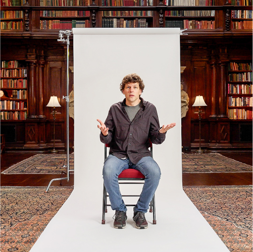 A man sits on a chair in front of a white backdrop, with bookshelves and lamps in the background, raising his hands in a questioning gesture.