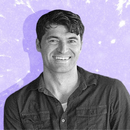 A smiling man with short dark hair wears a button-up shirt, standing in front of a purple, splattered-texture background.