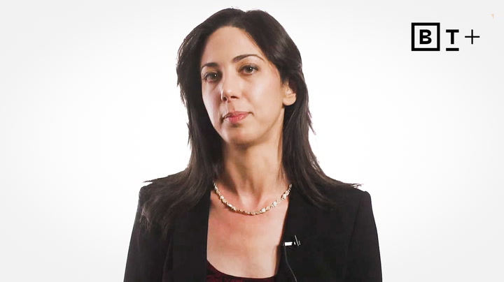 A woman with dark hair wearing a black blazer and necklace stands in front of a white background with a "B T +" logo in the top right corner.