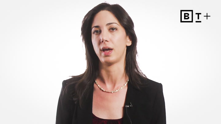 A woman with dark hair wearing a black blazer and necklace speaks in front of a plain white background with a "B T +" logo in the upper right corner.