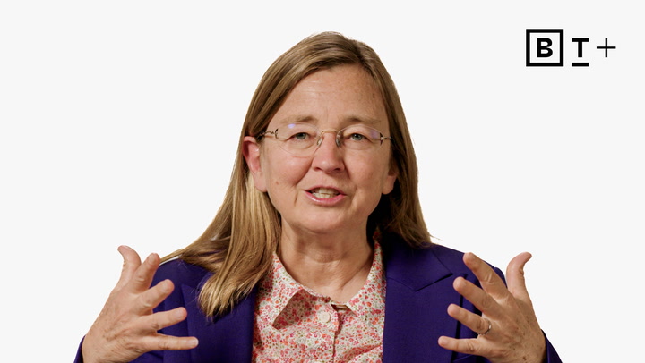 A woman with glasses and long light brown hair gestures with her hands while speaking; she wears a purple blazer over a floral shirt. A "BT+" logo appears in the top right corner.