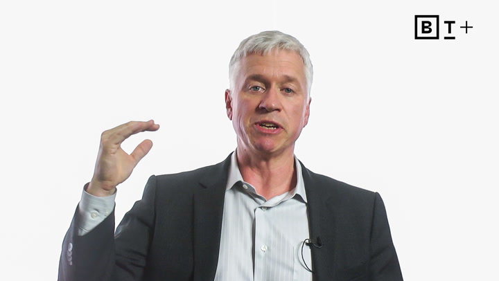 A man in a suit gestures with his right hand raised, while speaking. The background is plain white with a "BT+" logo in the top right corner.