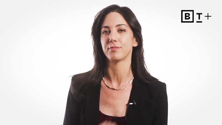 A woman with long dark hair, wearing a black blazer and necklace, sits in front of a plain white background with a "BT+" logo in the top right corner.