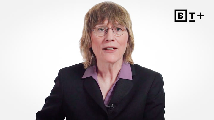 A person with shoulder-length light brown hair and glasses wearing a black blazer and purple shirt, seated in front of a white background with a "BT+" logo in the corner.