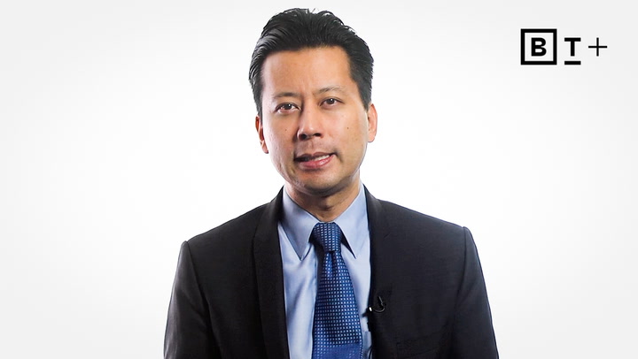 A man in a suit and blue tie stands against a plain white background with a "BT+" logo in the top right corner.
