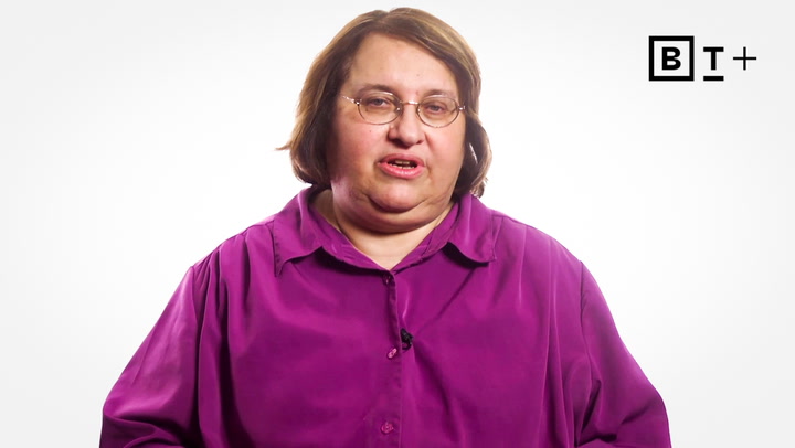 A person with shoulder-length brown hair and glasses wears a purple button-up shirt, sitting against a plain white background with a "B T +" logo in the top right corner.
