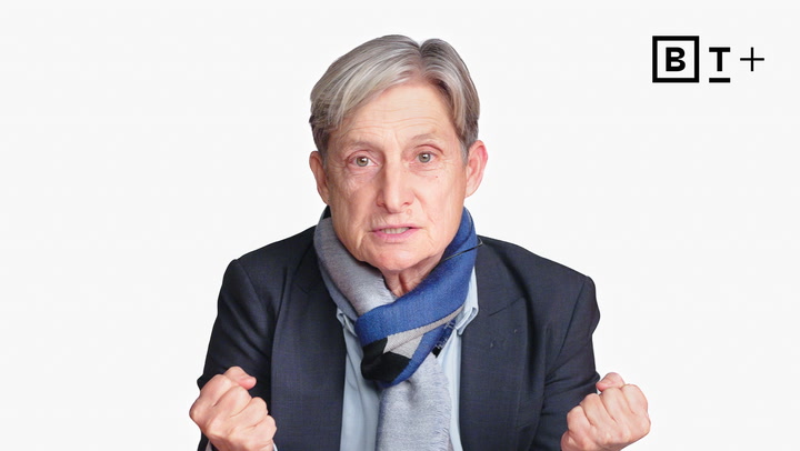 An older person with short gray hair and a blue scarf, dressed in a dark jacket, gestures with both hands. A black and white "B T +" logo is visible in the top right corner.