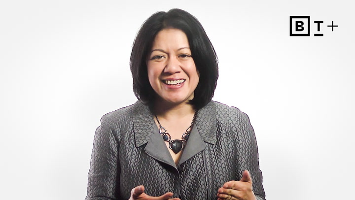 A woman with shoulder-length dark hair, wearing a patterned blazer and necklace, smiles and gestures while speaking in front of a white background with a "BT+" logo.