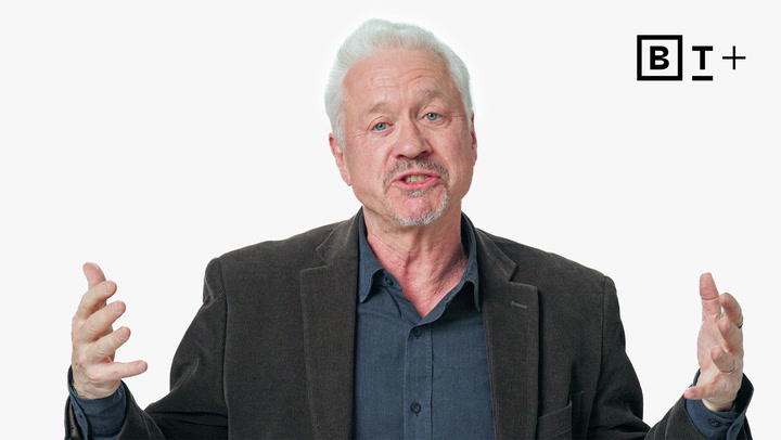 An older man with white hair and a goatee, wearing a dark blazer and shirt, gestures with his hands; a "BT+" logo is in the upper right corner against a plain white background.