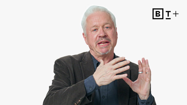 An older man with white hair, wearing a dark blazer and blue shirt, gestures with his hands while speaking in front of a white background featuring a "BT+" logo.