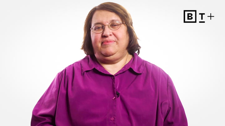 Person with shoulder-length brown hair and glasses wearing a purple shirt, standing against a plain white background with a "B T +" logo in the corner.