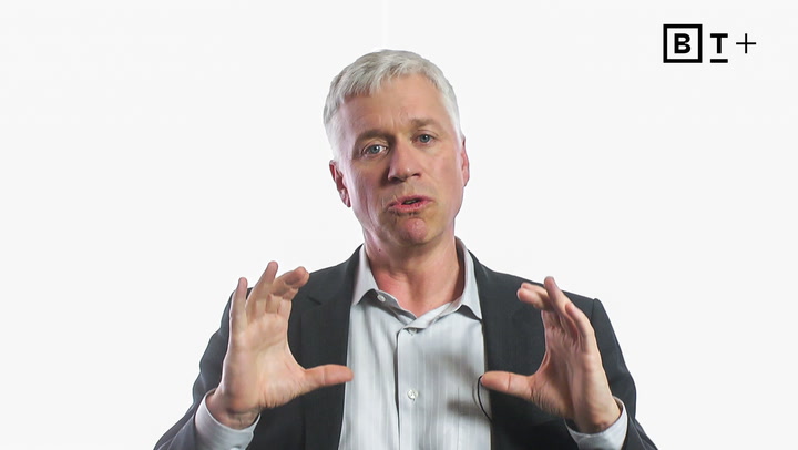 A man in a suit gestures with his hands while speaking in front of a plain white background with a "BT+" logo in the corner.