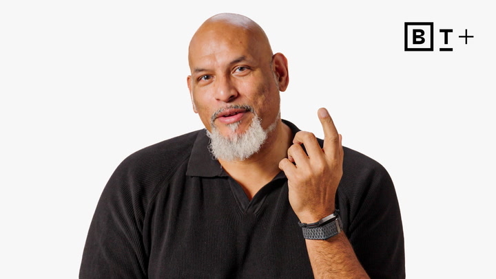 A bald man with a gray beard wearing a black shirt and smartwatch gestures with his right hand, with a "B T +" logo in the top right corner.