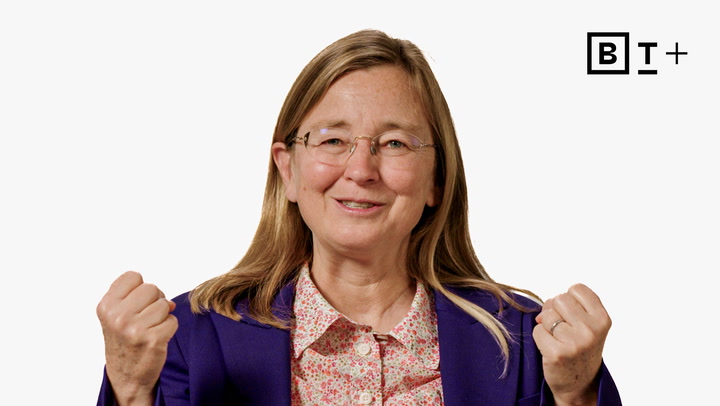 A woman with long light brown hair, wearing glasses and a purple blazer over a floral shirt, smiles and gestures with both fists raised. A "BT+" logo appears in the top right corner.
