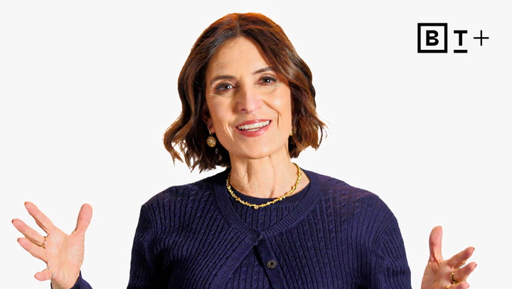 A woman with shoulder-length brown hair, wearing a navy sweater and gold jewelry, gestures with her hands against a plain white background with a "B T +" logo.